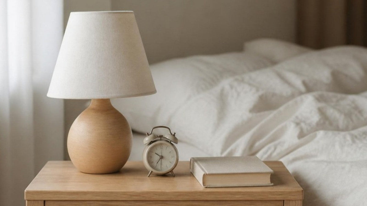 Lampe de chevet éteinte, réveil et livre fermé sur une table de nuit en bois près d'un lit aux draps de lin clair, article du Journal Calm&Co